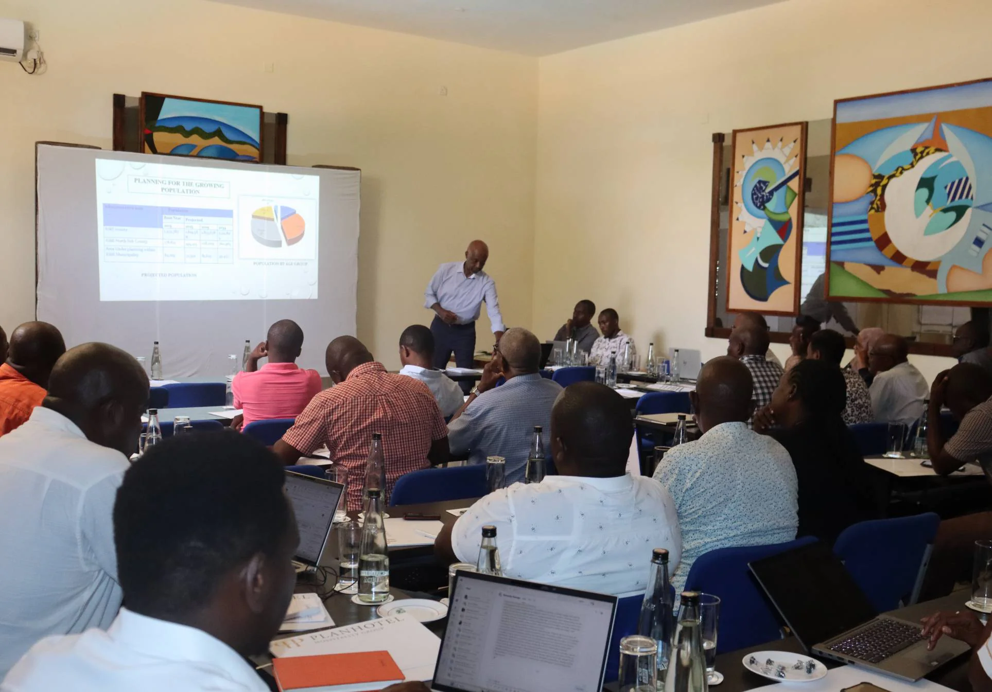 Participants seated at tables in a conference hall during a stakeholder engagement session, a presenter beside a projected slide