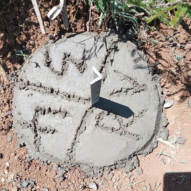 A newly cast concrete boundary beacon with an inscribed top and a steel marker, in red soil among dry grass