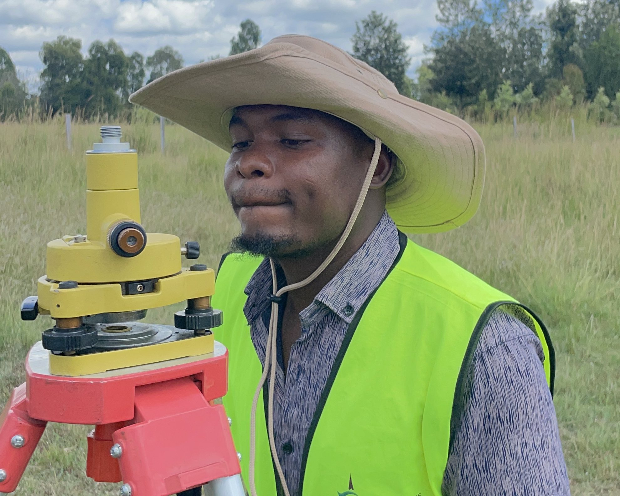 A GGIL surveyor in a company high-visibility vest and a wide-brimmed hat setting up a yellow tribrach on an orange tripod, sighting through its optical plummet, in open grassland