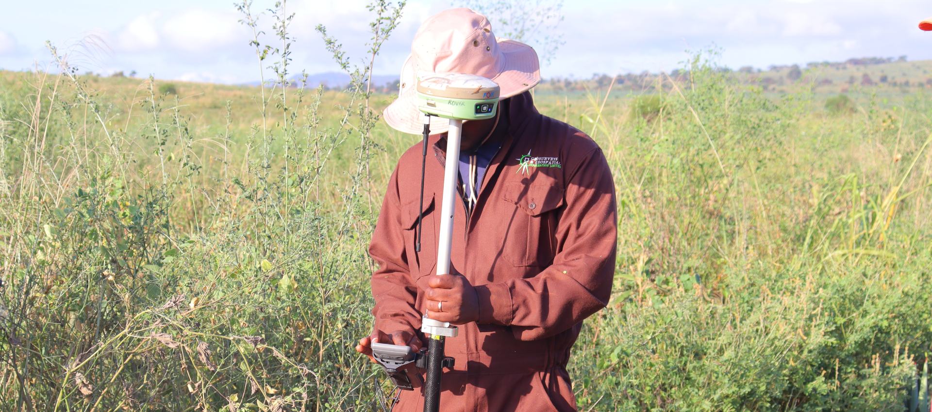 A GGIL field officer in a company jacket and a wide-brimmed hat holding a GNSS rover pole upright in tall grass, open hills behind