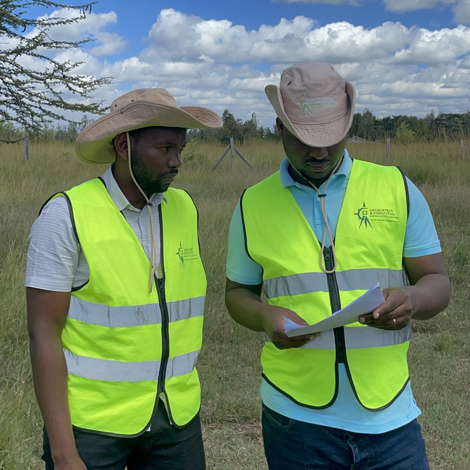 Two GGIL surveyors in company high-visibility vests and field hats reading a plan sheet together in open grassland, an acacia and a fence line behind them