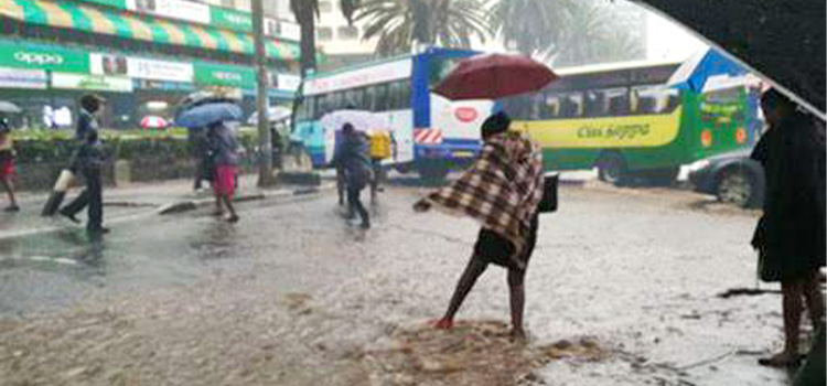 Pedestrians crossing a flooded city street in the rain, one wrapped in a checked blanket and others under umbrellas, with buses behind