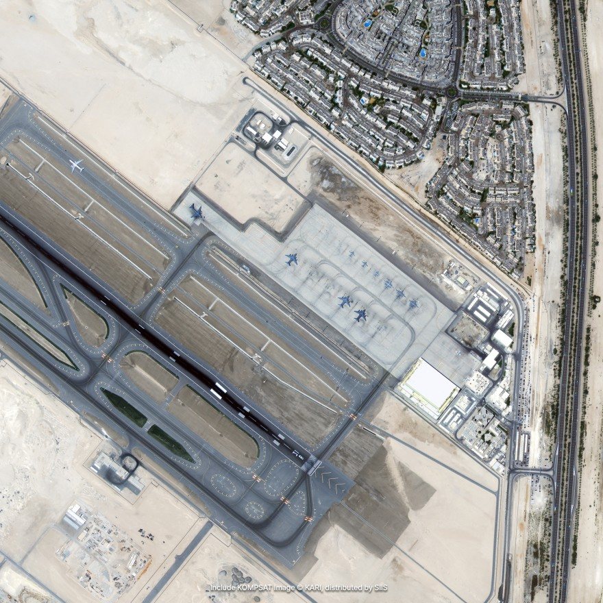 Satellite imagery of an airport runway and taxiways in desert, with aircraft on the apron and a town beside it