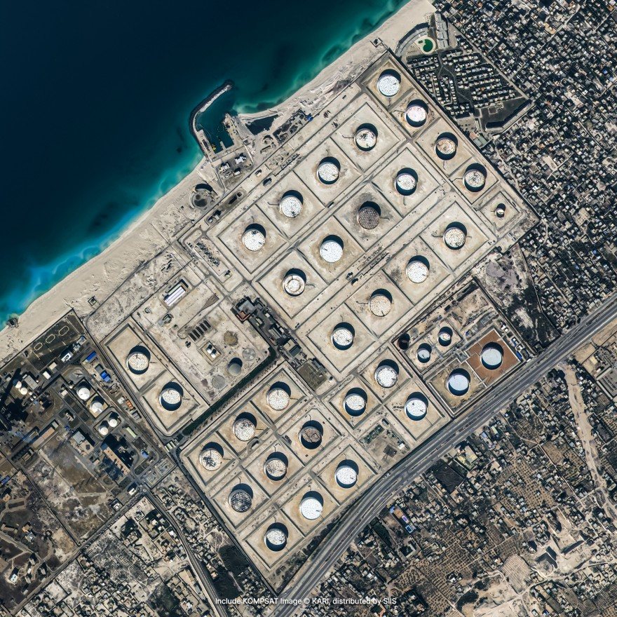Satellite imagery of an oil storage depot on a coast: rows of round storage tanks inside walled bunds, between the sea and a town