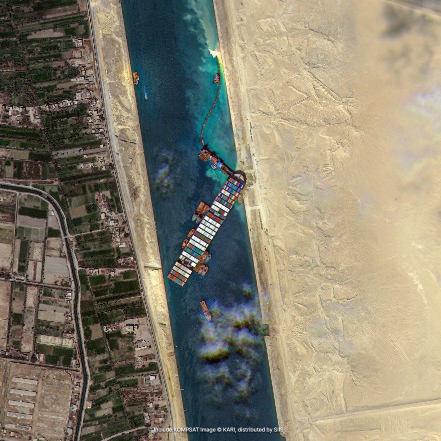 Satellite imagery of a large container ship lying across a narrow canal, with tugs beside it, between irrigated farmland and desert