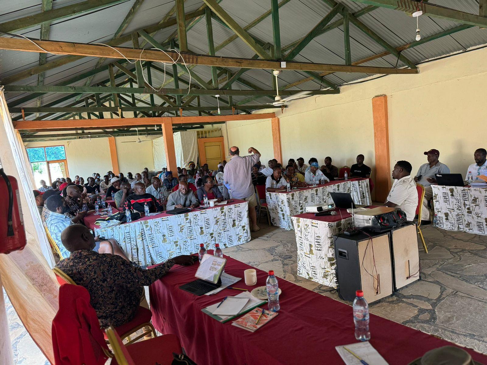 Participants seated at tables in a hall during a validation workshop, one standing to speak