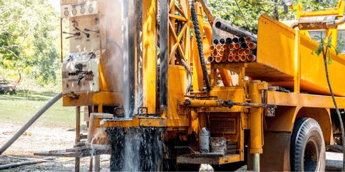 A yellow truck-mounted drilling rig at a borehole, water gushing from the casing